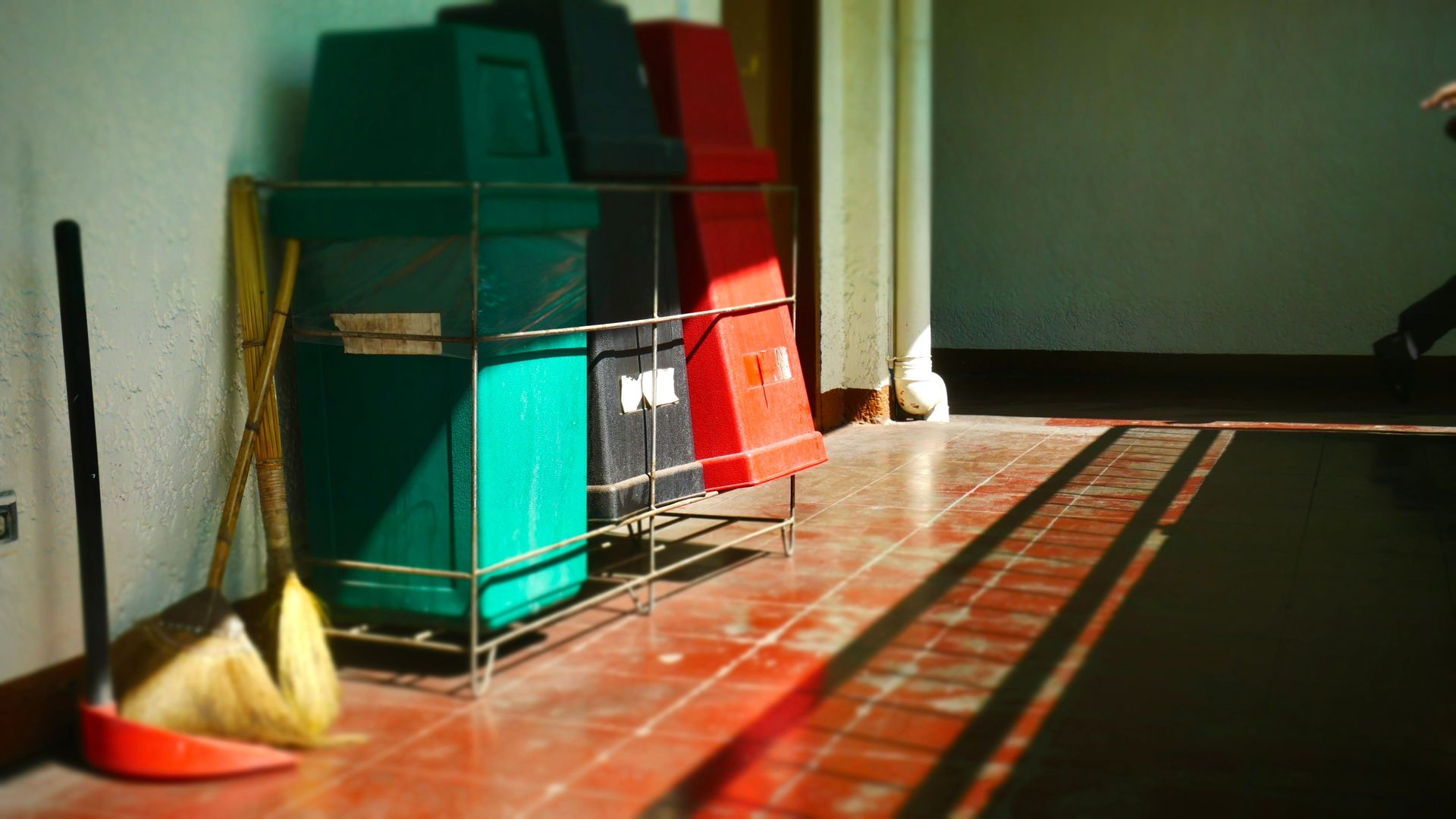 Trash bins and cleaning tools in a bright indoor corridor casting long shadows.