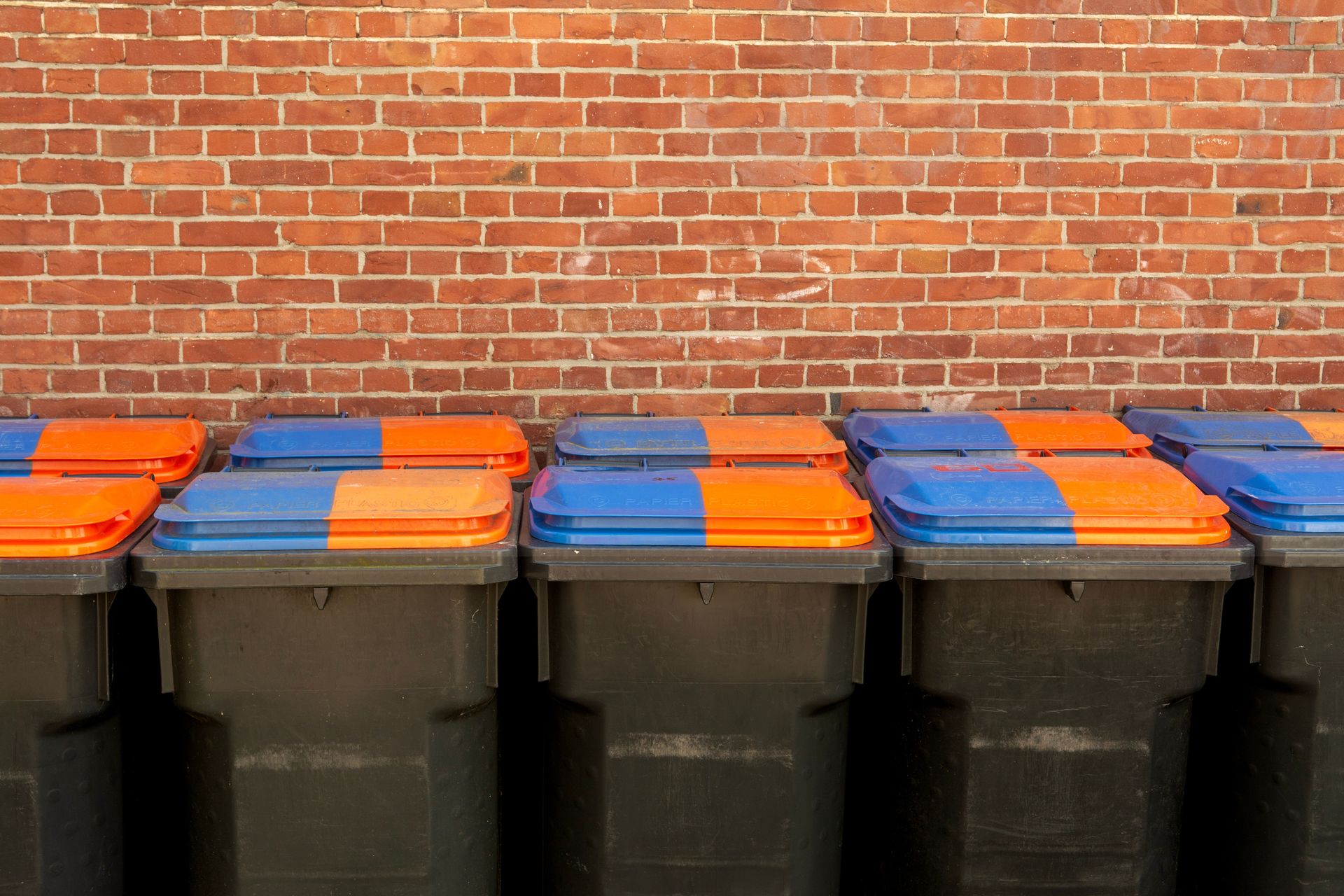 Row of recycling bins with vibrant lids in front of a brick wall for urban waste management.