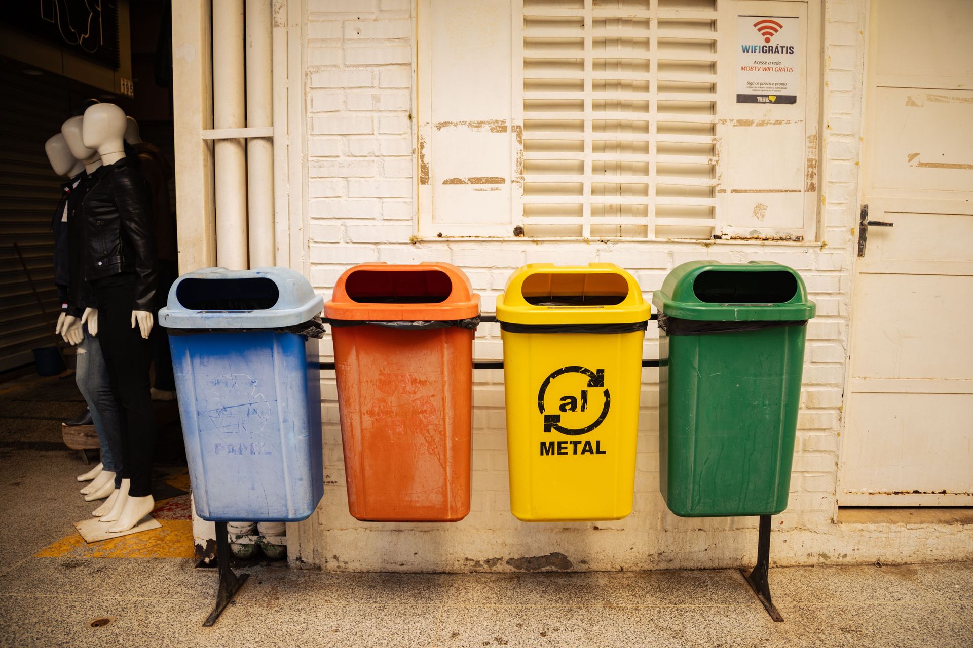 Four colorful recycling bins for different materials against a brick wall in an urban environment.