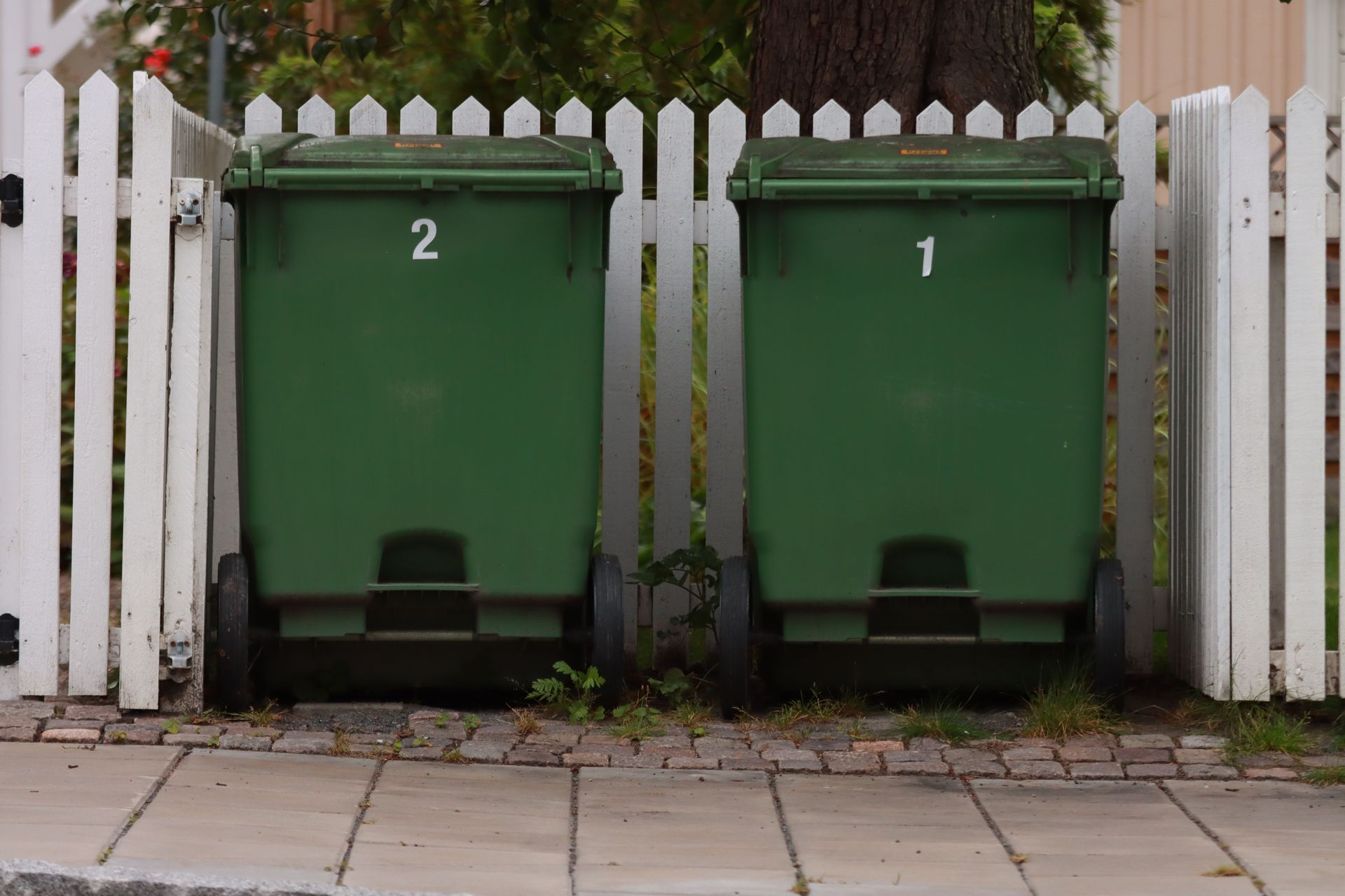 Two green garbage bins numbered 1 and 2 beside a white picket fence in Jönköping, Sweden.
