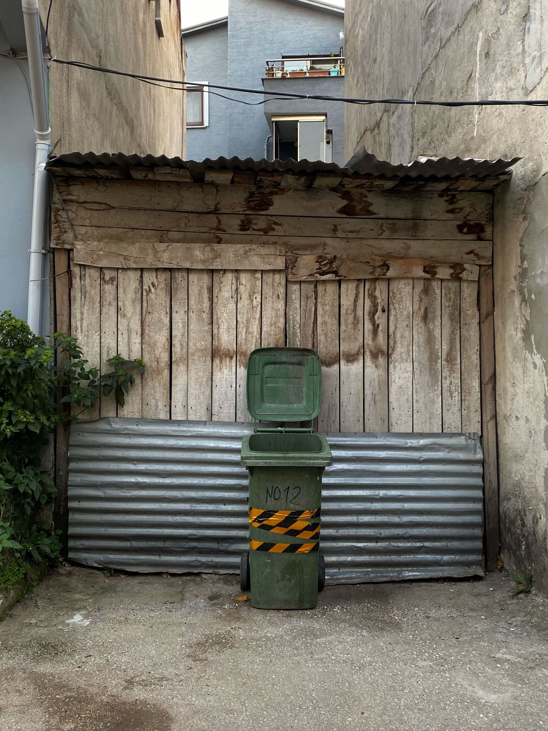 A rustic alley with a weathered wooden shed and a green trash bin in front.